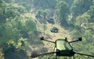 A green anti-tank loitering munition Rotem Alpha drone flies over a dense, sunlit forest with several military vehicles partially visible on a dirt clearing below, surrounded by trees and foliage.