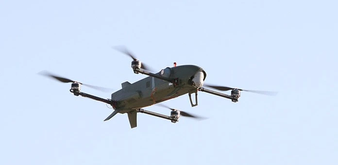 A gray quadcopter drone with four propellers, resembling a switchblade drone, is flying against a clear blue sky.