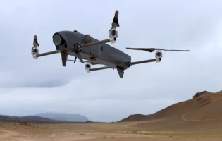 A gray IAI Rotem quadcopter drone with extended arms and propellers flies over a barren, rocky desert landscape under a cloudy sky.