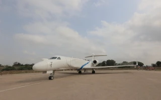 A white IAI jet aircraft with a blue stripe is parked on a concrete runway under a partly cloudy sky, with trees and open land in the background.