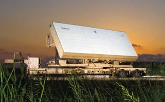 A large, rectangular military radar unit supporting network centric operations is mounted on a trailer in a grassy field at sunset, with a clear sky and the Lockheed Martin logo visible on the radar panel.