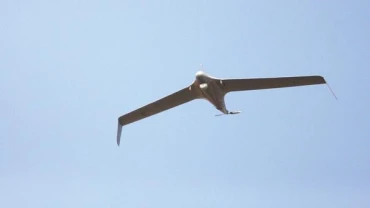 A drone with long, narrow wings is flying against a clear blue sky, viewed from below.