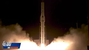 A rocket launches at night, surrounded by bright flames and smoke. The dramatic scene features the IAI logo and the words “In Action” in the lower-left corner as the rocket rises vertically into the sky.