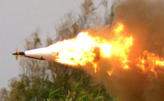 A missile is in flight, trailing bright flames and thick smoke against a backdrop of blurred greenery and cloudy sky.
