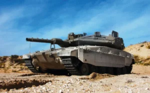 A modern IAI military tank with camouflage paint and a large cannon sits on rocky terrain under a clear blue sky, facing left. Desert vegetation and sandy hills are visible in the background.