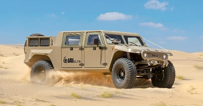 A tan, military-style off-road vehicle with large tires drives through a sandy desert, kicking up dust under a clear blue sky. The IAI-branded vehicle features ELTA and Zibar logos on its side.