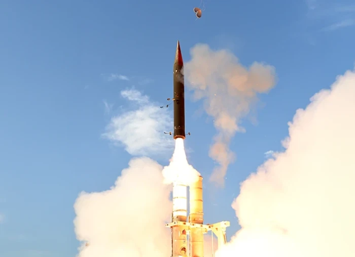 A missile is launching vertically into a clear blue sky, surrounded by clouds of smoke and flames at its base, with a small object visible above the missile.