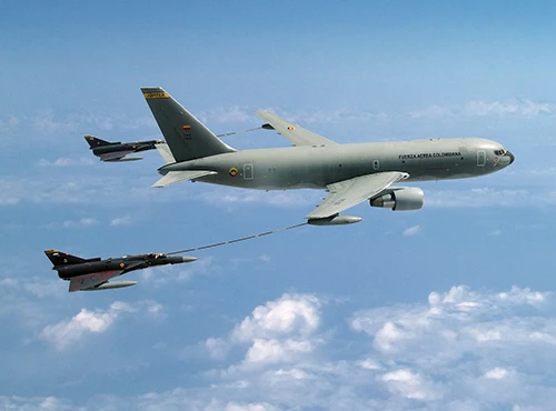 A Colombian Air Force tanker aircraft refuels two fighter jets mid-flight above the clouds during daylight. The tanker extends a hose to one jet while the other follows closely behind.