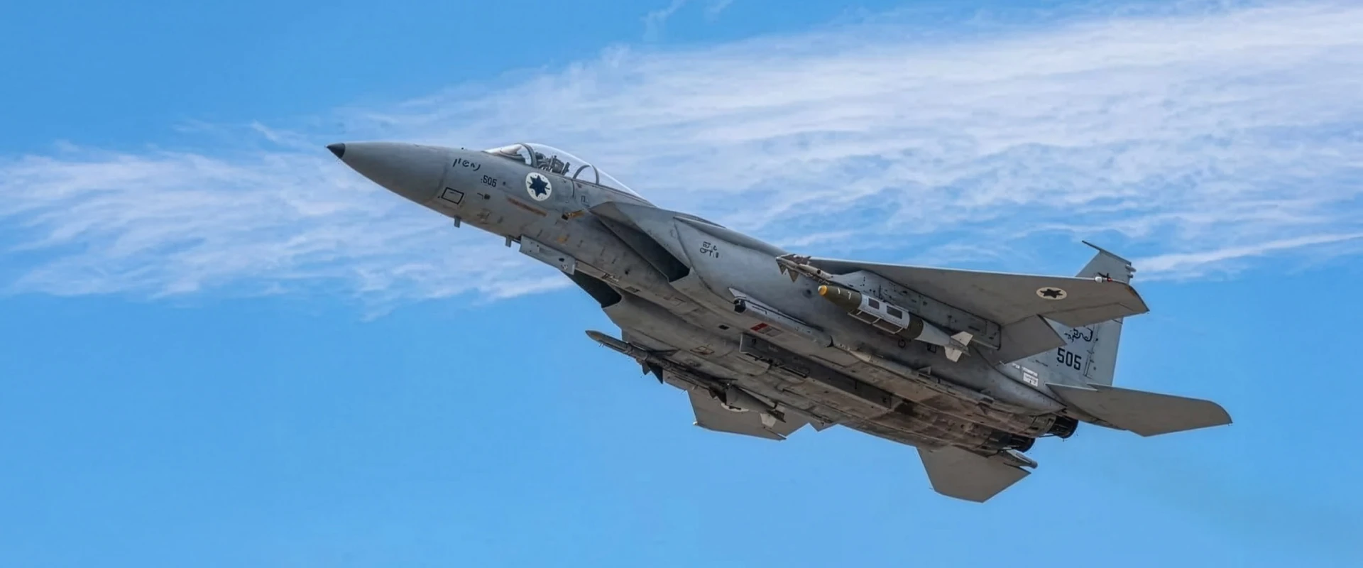 A military fighter jet with an Israeli Air Force insignia soars in a clear blue sky, its fuselage sections gleaming against the backdrop of wispy white clouds.
