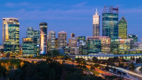 A vibrant city skyline at dusk, featuring modern high-rise buildings with lights on, set against a blue sky. Roads with moving vehicles and green trees are visible in the foreground.