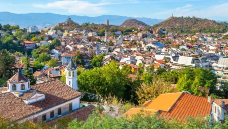 A panoramic view of a city with red-roofed buildings, green trees, and rolling hills in the background under a clear blue sky.