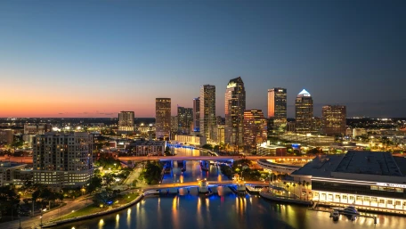 Downtown city skyline at dusk with illuminated buildings, a river with bridges reflecting lights, and a colorful sunset sky in the background.
