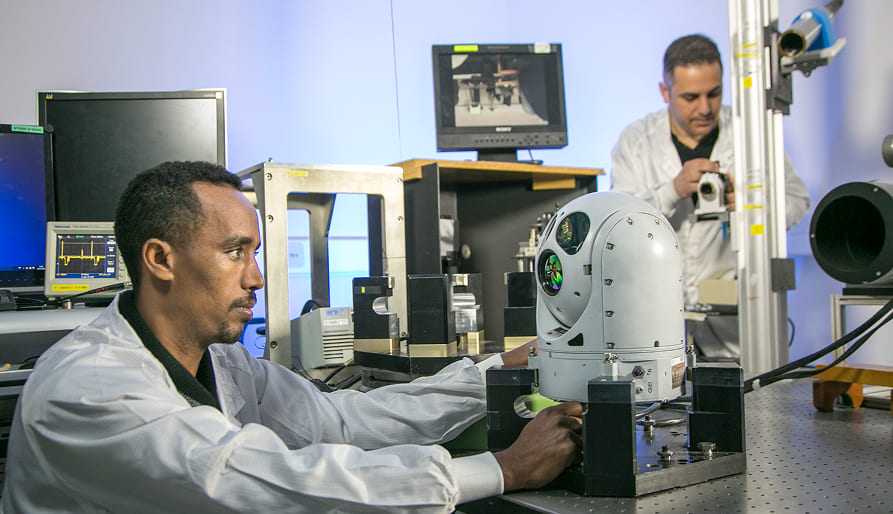 Two scientists in lab coats work in a laboratory with electronic equipment; one adjusts a spherical device on a table while the other operates machinery in the background, surrounded by monitors and technical instruments.