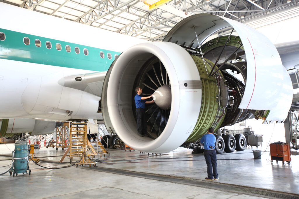 Two mechanics inspect and work on a large jet engine attached to a commercial airplane inside an aircraft hangar, surrounded by maintenance equipment and tools.
