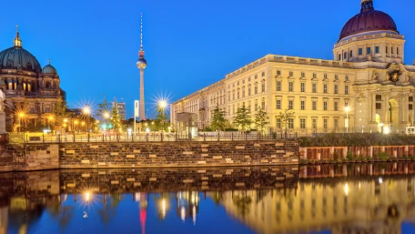 A riverfront view of Berlin at dusk, showing historic buildings, the Berlin TV Tower, and soft city lights reflecting in the calm water under a clear blue sky.