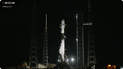 A rocket stands upright on a launch pad at night, illuminated by bright lights, with support structures and visible vapor emanating from its side. A countdown timer reads T - 00:07:49 in the top left corner.