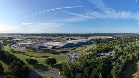 A panoramic view of an airport surrounded by greenery, with modern curved-roof buildings, open roads, parked vehicles, and a clear blue sky with jet trails overhead.