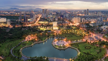A cityscape at dusk features a well-lit urban park with a large pond in the foreground, surrounded by greenery, and a sprawling city with tall buildings in the background.