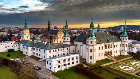 Aerial view of a large historic building complex with green-roofed towers and white walls, set against a cityscape at sunset with dramatic clouds in the sky.