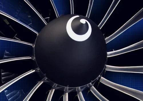 Close-up view of a jet engine fan, showing shiny metallic blades radiating from a dark circular hub with a white swirl design at the center. A blue grid background is partially visible behind the engine.
