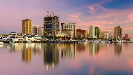 A city skyline with high-rise buildings under construction, waterfront structures, and boats docked along a calm bay at sunset. The sky is colorful and the buildings are reflected in the water.