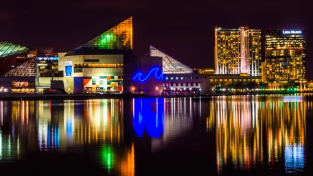 Baltimore’s Inner Harbor at night, featuring the illuminated National Aquarium and nearby buildings reflected in the calm water, with colorful lights creating vibrant reflections.