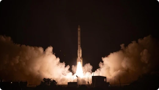 A rocket launches into the night sky, surrounded by clouds of smoke and bright flames at its base, illuminating the darkness around the launch site.